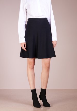 Femme portant une jupe noire au genou, une chemise blanche à manches longues et des bottines noires à talons, debout devant un fond uni.