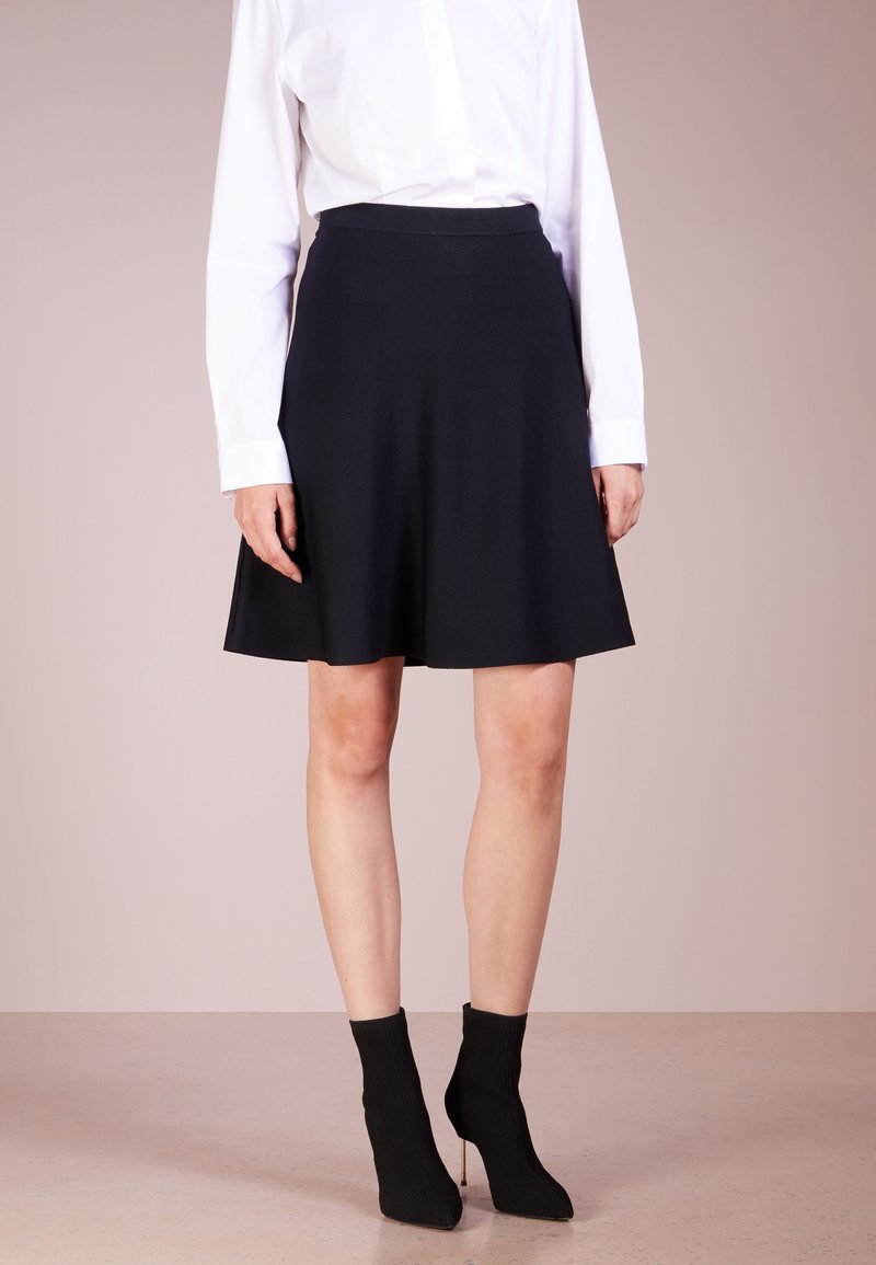 Femme portant une jupe noire au genou, une chemise blanche à manches longues et des bottines noires à talons, debout devant un fond uni.