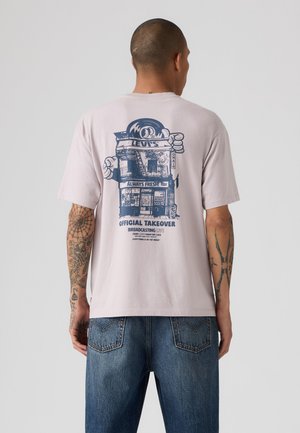 Homme avec des tatouages portant un T-shirt rose clair avec un motif bleu représentant un bâtiment et un texte dans le dos, associé à un jean bleu, vu de dos.