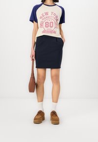 T-shirt med raglanärmar i krämfärgad och marinblå, med tryck av "NEW YORK", parkerad med en åtsittande marinblå kjol. Brun axelväska och platåsandaler.