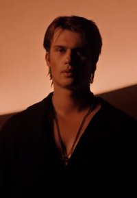 Jonge man met kort haar draagt donkere kleding en gelaagde kettingen, verlicht door warm zacht licht met een wazige achtergrond.