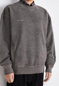 Sweat-shirt gris avec une finition texturée, des poignets et un ourlet côtelés. Présente un logo "HUGO" brodé sur la poitrine et des poches latérales.
