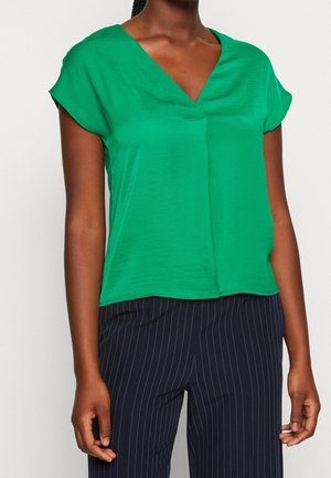 Frau trägt eine leuchtend grüne kurzärmelige V-Ausschnitt-Bluse kombiniert mit marineblauen Nadelstreifen-Hosen und steht vor einem einfarbigen Hintergrund.