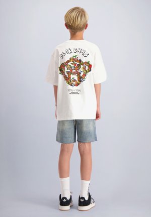 Kind met kort blond haar, gekleed in een wit T-shirt met print, spijkershorts, witte sokken en zwarte Adidas sneakers, staand met de rug naar voren.