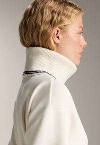 Persoană blondă văzută în profil, îmbrăcată cu un pulover alb cu guler înalt și pliuri, având o dungi negre aproape de marginea gulerului.