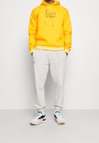 Sudadera amarilla con capucha y cordones, rayas blancas en los puños; combinada con pantalones de chándal gris claro con texto sutil. Zapatillas blancas con detalles.