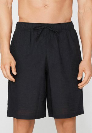 Man draagt zwarte knielange korte broek met elastische taille en trekkoord, toont de buik en handen aan de zijkanten.