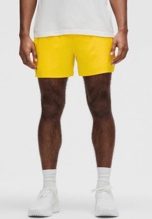 Gelbe Shorts aus Baumwollstoff mit glatter Textur, die ein einfaches Design und einen elastischen Bund aufweisen. Getragen mit weißen Turnschuhen.