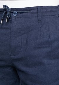 Granatowe spodnie z teksturowanej tkaniny, z elastyczną talią i zapięciem na guzik. Posiadają kieszenie z przodu oraz plisowane detale w projekcie.