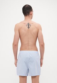 Modri in beli črtasti kopalni šortki z elastičnim pasom, ki kažejo gladko teksturo. Dekorativna tetovaža je vidna na zgornjem delu hrbta.