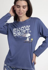 Camiseta de pijama de manga larga azul con el texto blanco "SNOW DRIFT" y un gráfico de Snoopy y Woodstock. Tela suave y corte relajado, con costuras en contraste.