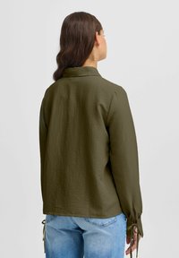 Olive green, long-sleeve shirt with a textured surface. Ties at the cuff and a collar, paired with light blue jeans. Simple design and silhouette.