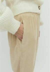 Beige Cordhosen mit gerippter Textur, versehen mit Seitentaschen und einem hohen Bund, kombiniert mit einem weichen, weißen Strickpullover.