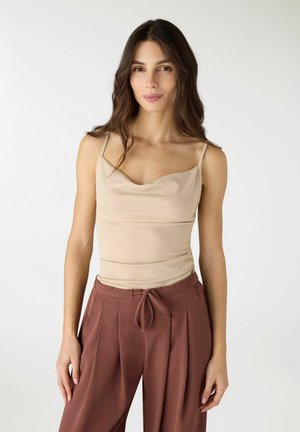 Donna con capelli lunghi e scuri che indossa un top beige senza maniche e pantaloni marroni a vita alta legati con un fiocco, in piedi davanti a uno sfondo bianco.