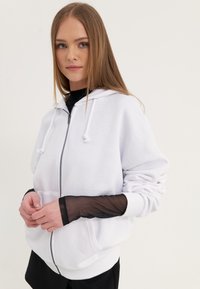 Biela mikina na zips s kapucňou a šnúrkami, so jemnou textúrou a voľným strihom. Dlhé čierne sieťované rukávy poskytujú kontrast a detail.