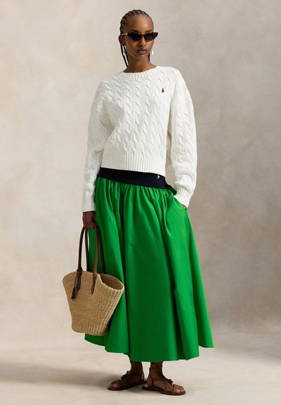Πουλόβερ κρεμ με πλέξη καλώδιο, ζωντανή πράσινη μίντι φούστα, καφέ σανδάλια και πλεκτή ψάθινη τσάντα, σε ουδέτερο φόντο.