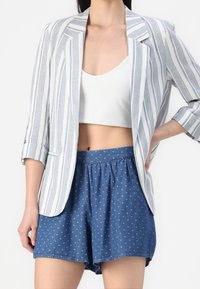 Žena v bílém crop topu, modrých šortkách s malými bílými diamantovými vzory a bílém blejzru s vertikálními modrými pruhy, ruka na boku.