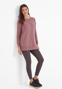 Light mauve sweatshirt with long sleeves, ribbed hem, and side slits, paired with dark grey leggings and black athletic shoes.