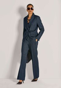 Vrouw in een donker streepjassenpak met een dubbelgeknoopte blazer, brede broekspijpen, zwarte hoge hakken, grote zonnebril en ring oorbellen.