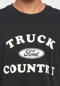 Fekete pamut póló, amelyen egy nagy fehér mintázat található, rajta a "TRUCK" és "COUNTRY" feliratokkal, a közepén egy fehér ovális Ford logóval.