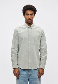 Svetlo zelena srajca z gumbi iz teksturirane tkanine z žepom na prsih, v kombinaciji s svetlo modrimi kavbojkami. Ponaša se z zaobljenim robom in ovratnikom z gumbi.