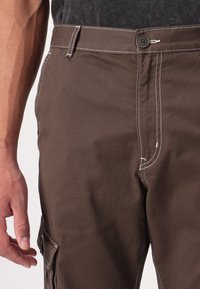 Brown cotton cargo pants with white stitching, featuring a front pocket and button closure, shown from the waist to mid-thigh.