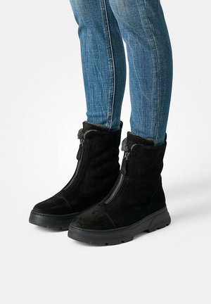 Schwarze Wildleder-Stiefeletten mit Reißverschluss, ausgestattet mit einem weichen, pelzigen Kragen und einer dicken schwarzen Gummisohle, kombiniert mit blauen Skinny-Jeans.