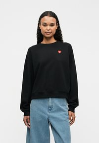 Svart hettegenser laget av bomull, med rund hals og dropped shoulders. Den har en liten rød hjerte-logo på brystet.