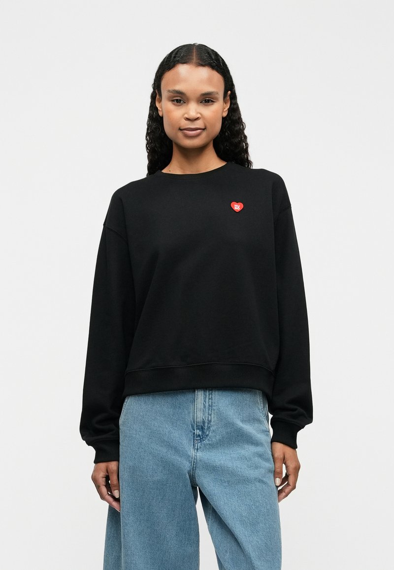 Svart hettegenser laget av bomull, med rund hals og dropped shoulders. Den har en liten rød hjerte-logo på brystet.