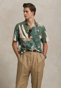 Korte mouwen groene shirt met zeilbootprint, gecombineerd met beige geplooide broek. Het shirt heeft een knoopkraag en een pocketdetail.