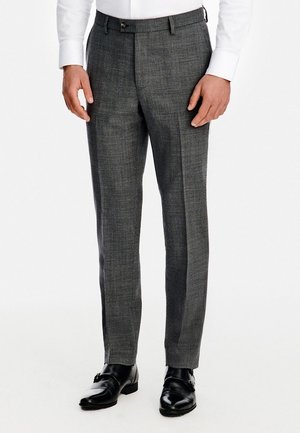 Mężczyzna ubrany w szare eleganckie spodnie, czarne skórzane buty wizytowe oraz białą koszulę z długim rękawem, stoi na tle jednolitego białego tła.