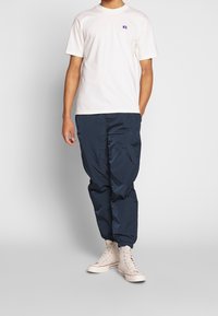 Man i vit t-shirt med kort ärm, marinblå nylonjoggingbyxor och off-white högstövlar står mot en neutral bakgrund.