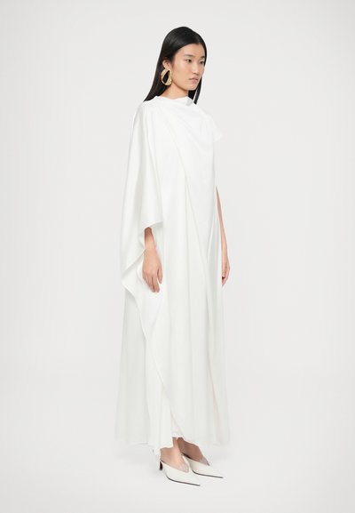 Bijela, lepršava haljina s naboranim dizajnom i širokim rukavima, izrađena od meke tkanine. Uparena s bijelim cipelama šiljastih nosa i velikim zlatnim naušnicama.