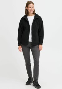 Veste en polaire noire avec une fermeture éclair à l'avant, un col haut et une surface texturée, associée à un pantalon gris slim-fit et des chaussures de sport noires.