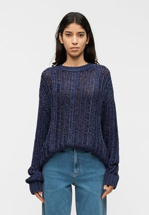 Løs, marineblå strikket sweater med vide ærmer, tekstureret åben vævning og ribbede manchetter, kombineret med lyseblå jeans.