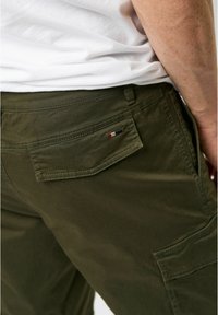 Pantalonii cargo de culoare verde măslin au un buzunar pe spate cu o etichetă mică. Materialul este neted, cu cusături vizibile și o croială lejeră.