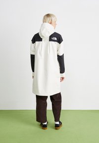 Manteau résistant à l'eau de couleur crème avec des accents noirs, capuche et logo. Stylé avec un pantalon en velours côtelé marron foncé et des chaussures noires. Design simple.