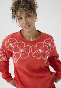 Femme aux cheveux bouclés portant de grandes boucles d'oreilles en or et un sweat-shirt rouge orné de grandes fleurs blanches brodées sur la poitrine et les manches.