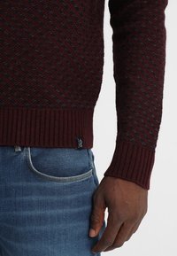 Burgunderfarbener Strickpullover mit diagonaler Struktur, gerippten Bündchen und einem kleinen Etikett am Saum, kombiniert mit hellblauen Denim-Jeans.