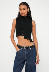 Μαύρο κοντό τοπ χωρίς μανίκια με ψηλό λαιμό, με ξεφτισμένα άκρα και μικρό λευκό λογότυπο. Φορεμένο με τζιν ψηλόμεσα και μαύρη ζώνη.