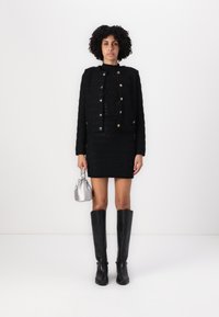 Sacou negru din tweed cu nasturi aurii, asortat cu o fustă mini neagră și cizme negre până la genunchi. Ținând o mică poșetă argintie.