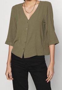 Femme portant une blouse olive à boutons avec des manches trois-quarts et un pantalon noir, accessoirisée d'un collier en chaîne dorée.