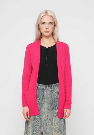 Ung kvinde med langt platinblond hår iført en lys pink cardigan, sort knap-top og grå slangeprint nederdel, stående foran en hvid baggrund.