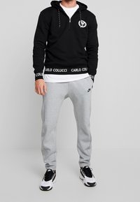 Felpa nera con zip e patch del logo, dotata di cappuccio regolabile e cintura con marchio; abbinata a pantaloni della tuta grigi chiari. Le sneaker completano il look.