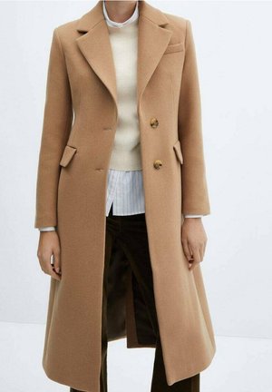 Manteau long en laine couleur beige avec revers à encoche et deux boutons, porté sur un pull blanc et une chemise rayée, associé à un pantalon foncé.