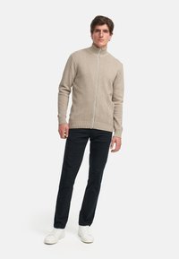 Suéter de punto beige con cremallera y patrón de chevron, cuello alto y cremallera blanca en contraste, combinado con pantalones oscuros y zapatillas blancas.