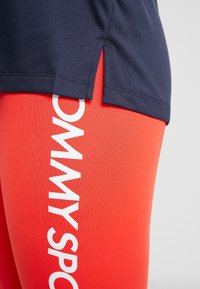 Marinblå idrottstopp med sidosprund, ihopparad med röda leggings som har vit "TOMMY SPORT" text vertikalt längs benet.