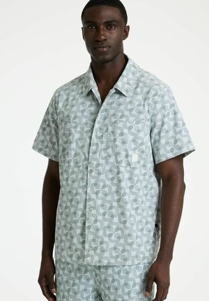 Mann trägt hellgraues, kurzärmeliges Hemd und passende Shorts mit dunklem geometrischem Wirbelmuster, steht vor weißem Hintergrund.