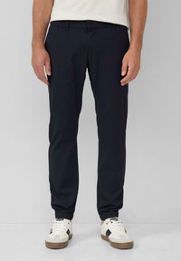 Marineblauwe broek met een elastische tailleband en taps toelopende pijpen, gemaakt van een gladde stof. In combinatie met witte en bruine sneakers.