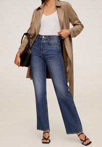 Jeansy z wysokim stanem, proste nogawki w kolorze niebieskim, w połączeniu z beżowym płaszczem typu trench. Stylizacja zawiera białą koszulkę oraz czarne sandały na paski.